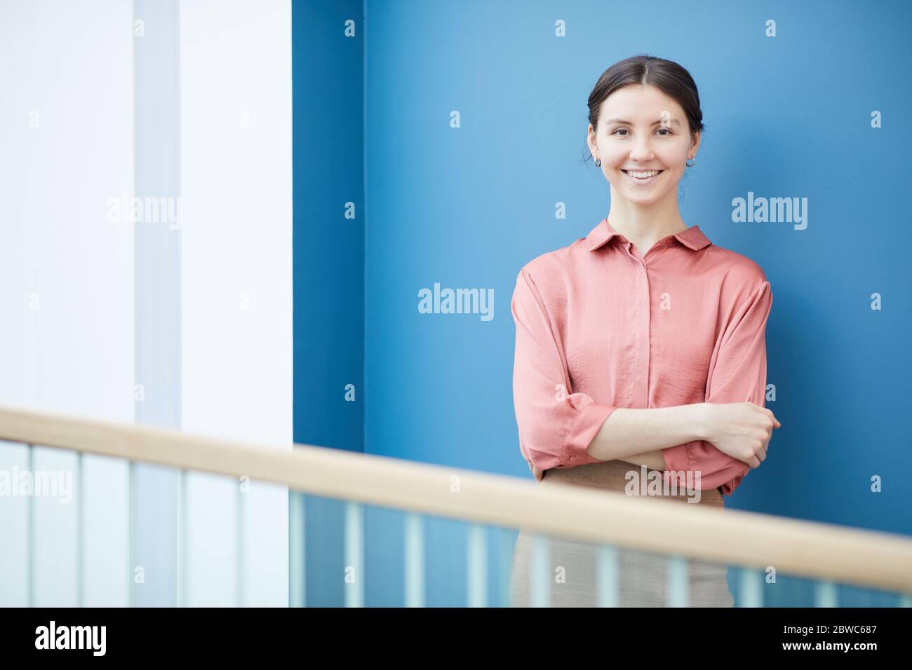 Waist up portrait of young elegant businesswoman smiling at camera while leaning against blue wall, copy space Stock Photo
