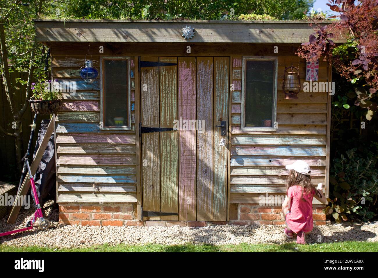 Young girl sat on floor colouring in shed wall in garden, Uk Stock Photo