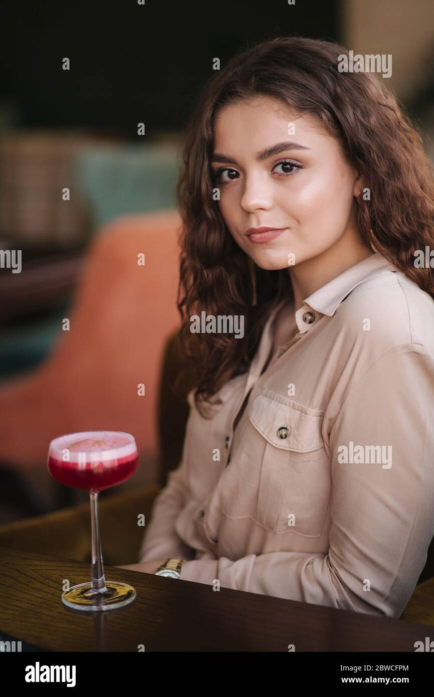 Young woman sitting in restaurant and drink cocktail. Beautiful girl with curly hair smile to camera. Smile Stock Photo