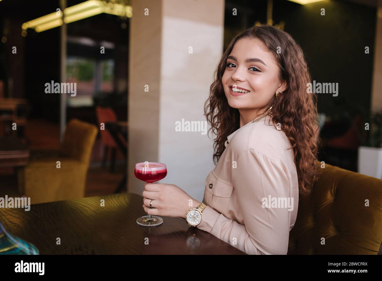 Young woman sitting in restaurant and drink cocktail. Beautiful girl with curly hair smile to camera Stock Photo
