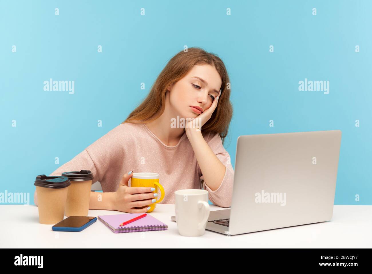 Exhausted bored woman, office employee surrounded by coffee cups in workplace, looking at laptop screen with indifferent apathetic face, overtime work Stock Photo