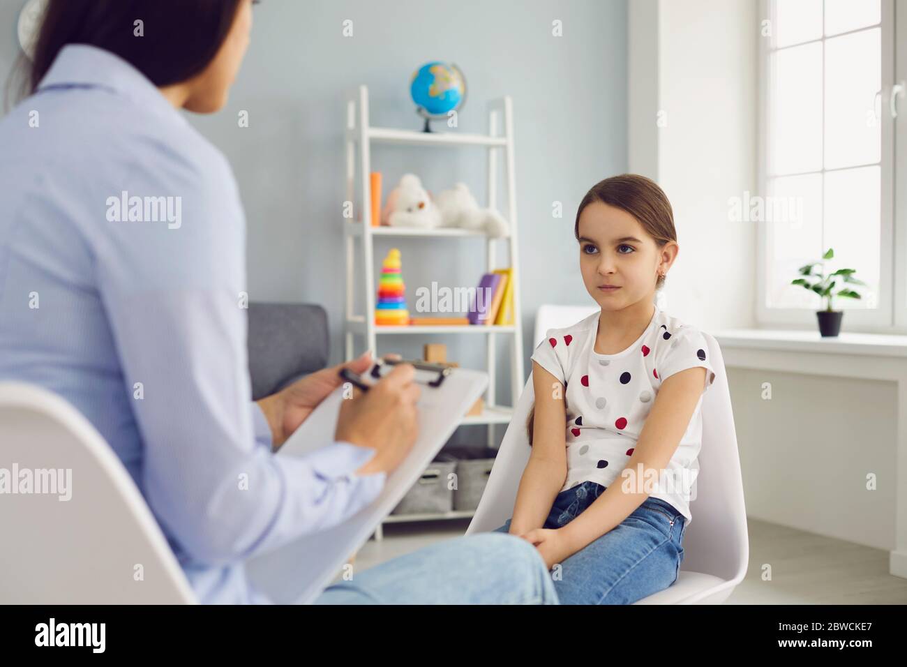 Young woman psychologist speaks with a child girl with problems sitting on a chair in the room. Stock Photo