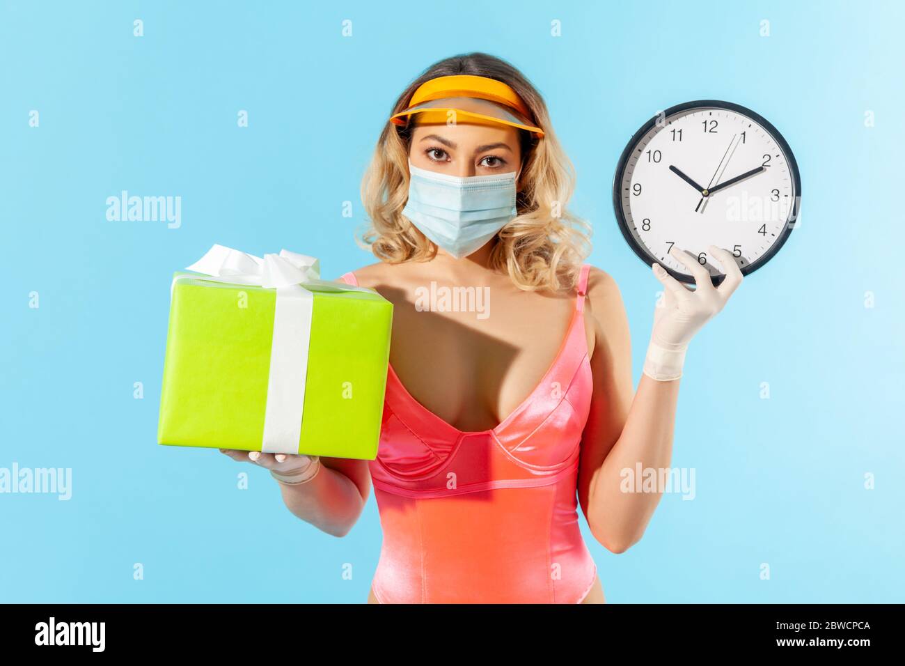 Birthday celebration, bonus in quarantine! Woman in swimsuit, hygienic face mask and protective gloves, holding gift box and big clock, safe summer ho Stock Photo