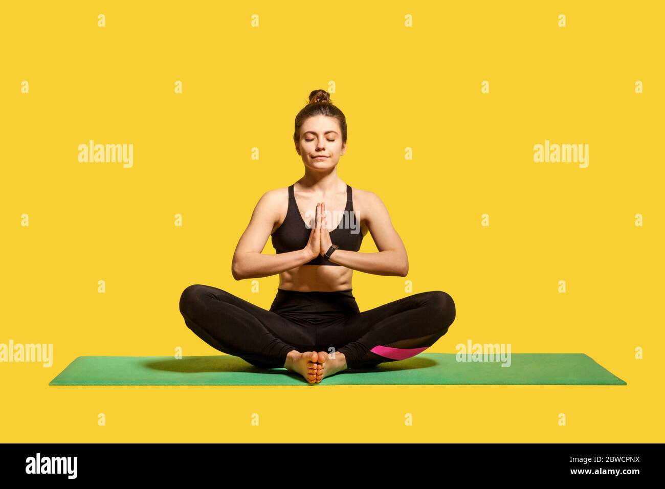 Concentrated peaceful woman with hair bun in tight sportswear sitting on mat practicing yoga, holding hands in namaste gesture and meditating, relaxin Stock Photo