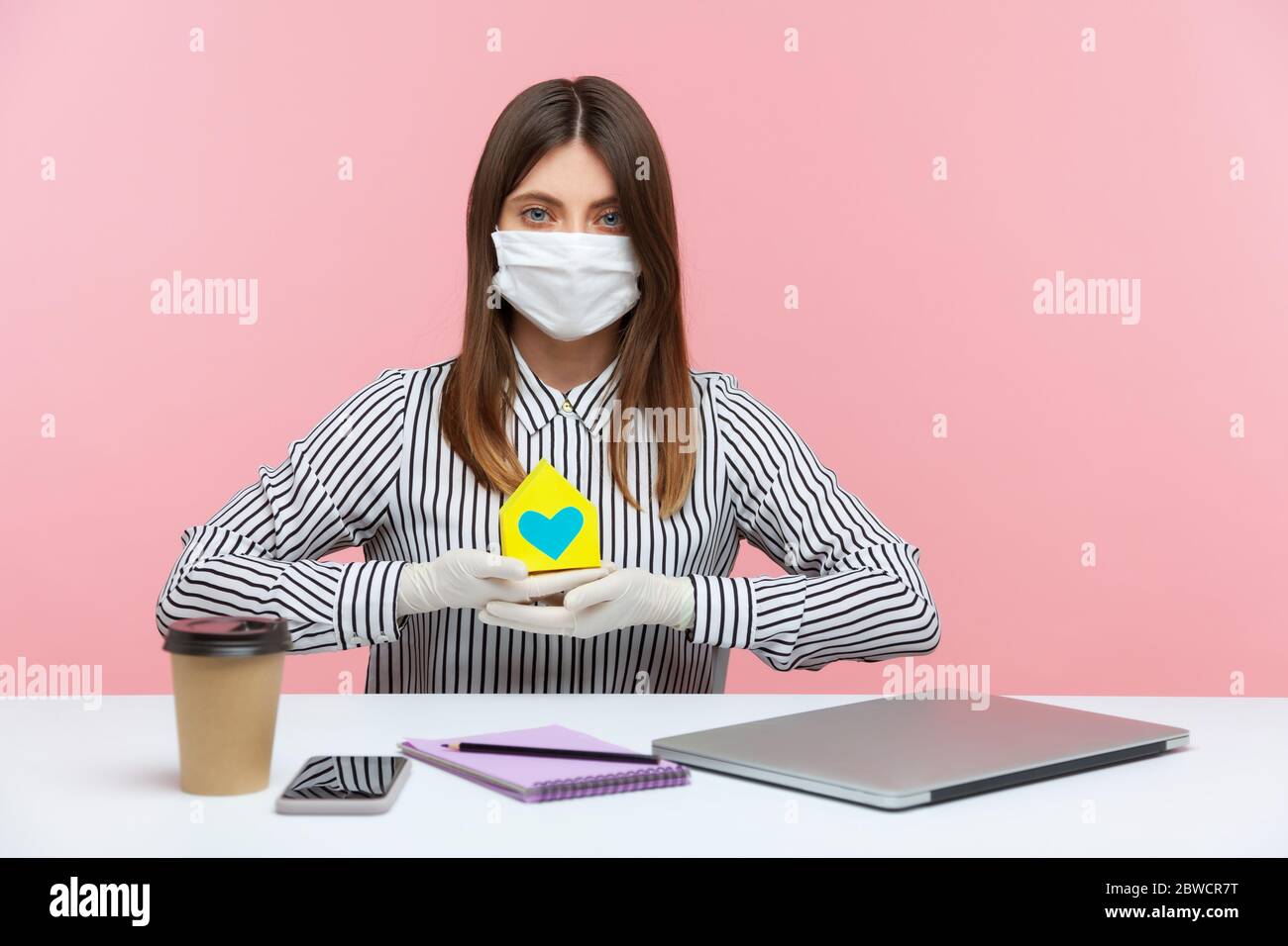 Renting during coronavirus quarantine. Woman real estate agency sitting at workplace, wearing hygienic mask and protective gloves, holding paper house Stock Photo