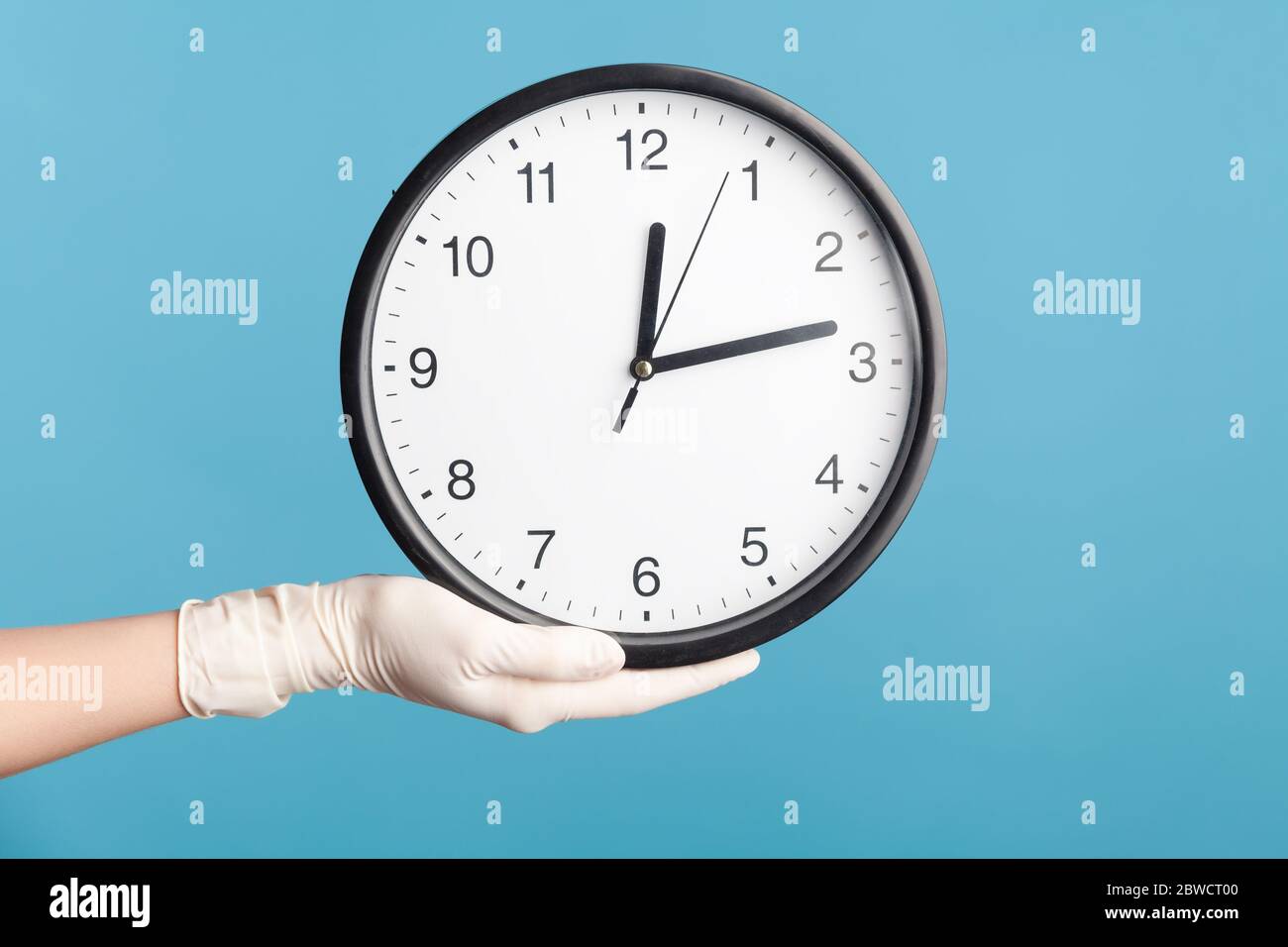 Profile side view closeup of human hand in white surgical gloves holding analog clock. indoor, studio shot, isolated on blue background. Stock Photo