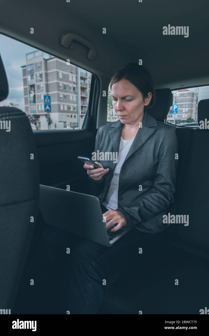 Businesswoman using mobile phone and laptop computer in car whole sitting at the back seat, business communication on the move Stock Photo