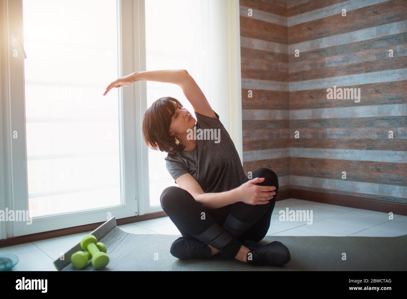 Adult fit slim woman has workout at home. Senior model sit with legs crossed and stretches arm to side. Exercising alone to get in good shape and Stock Photo