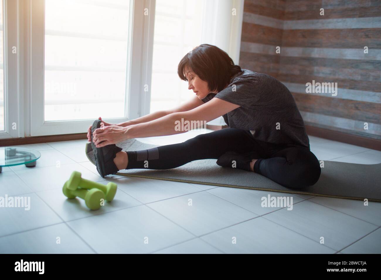 Adult fit slim woman has workout at home. Senior model sit with crossed legs and meditating or praying. Stretching hands holding together. Take care Stock Photo