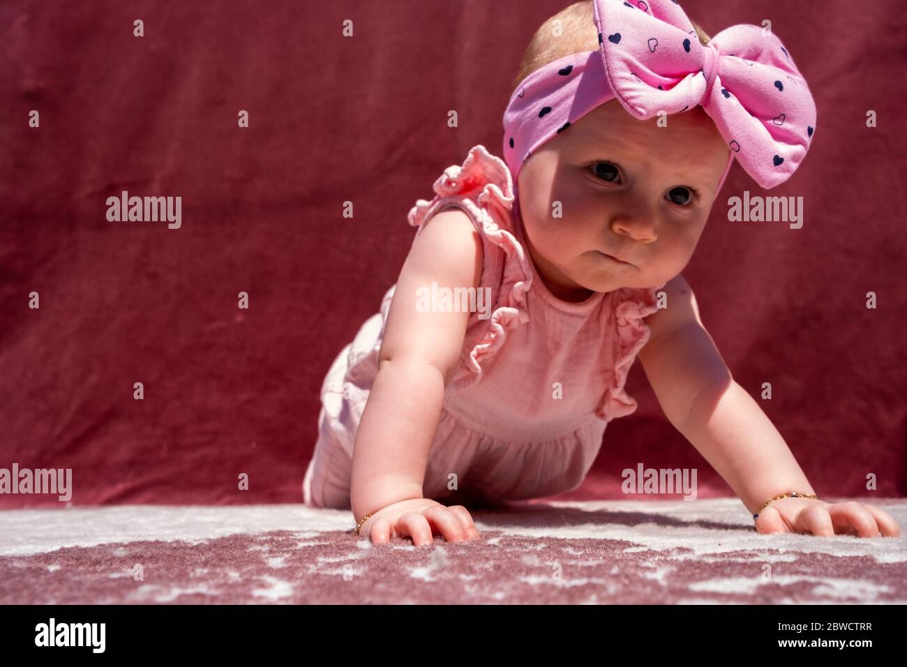 Cute baby girl wear adorable hat doing push-up outdoors, looking at camera and smiling on red background Stock Photo