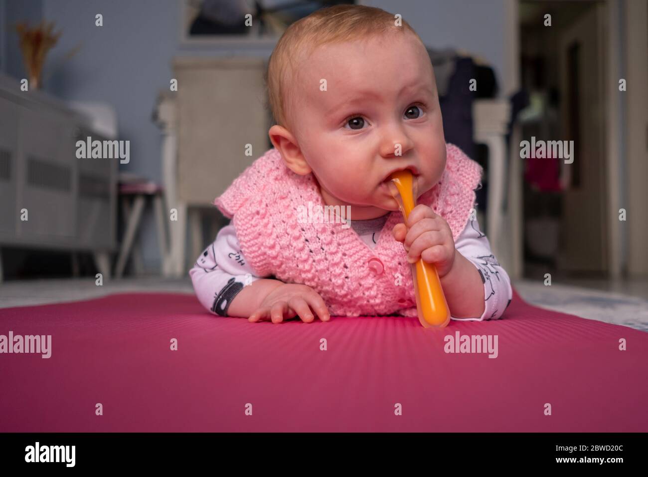 Cute baby girl lying on the pink yoga mat, holding and biting the spoon