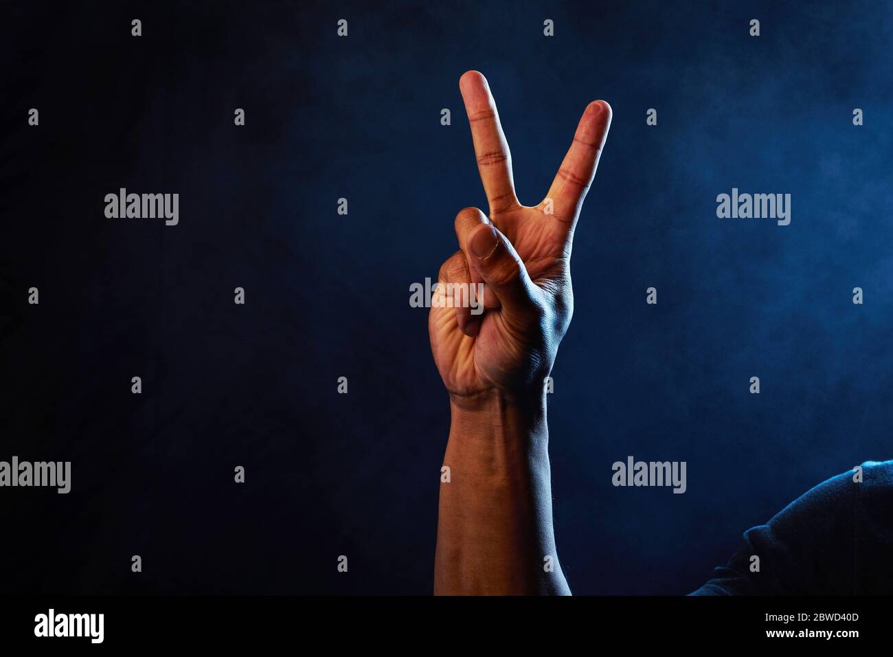 Studio image of an african american male hand holding up a peace sign with smoke background and blue lights representing police. Stock Photo