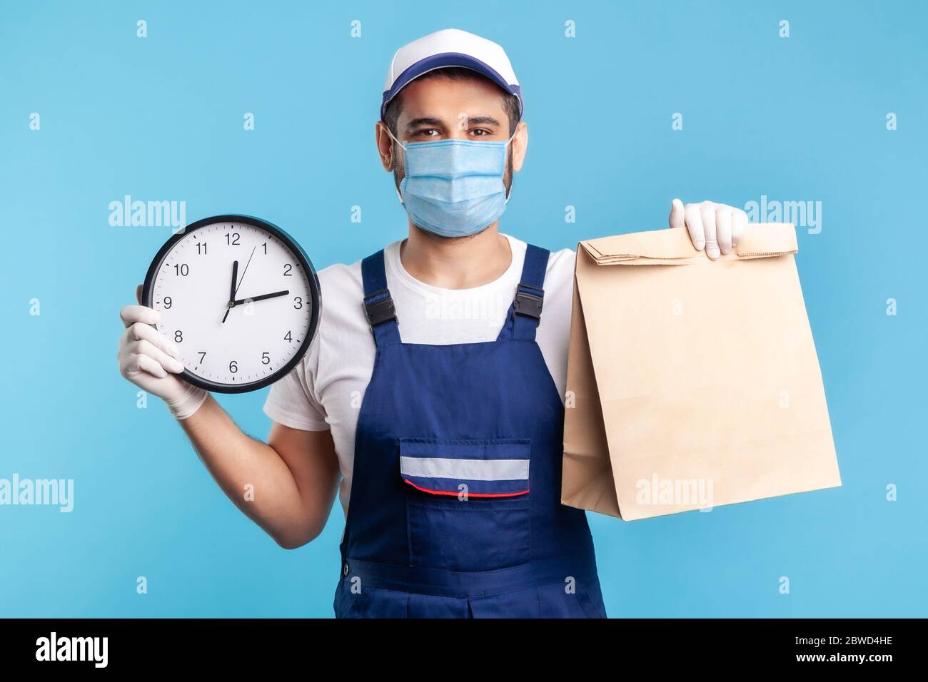 Food delivery, shipping on time! Portrait of handyman in uniform, mask, gloves holding clock and parcel. Express courier transportation, post mail ser Stock Photo