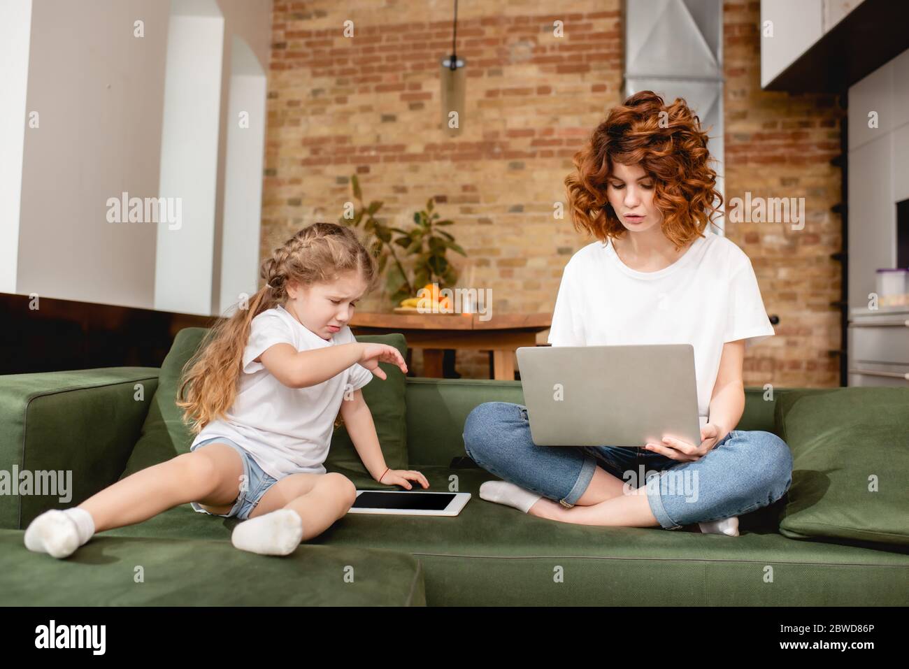 redhead freelancer using laptop near upset daughter with digital tablet Stock Photo