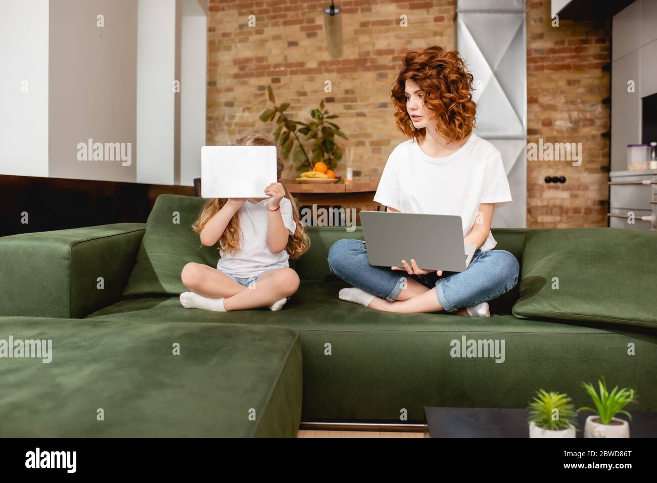 redhead freelancer using laptop near daughter covering face with digital tablet Stock Photo