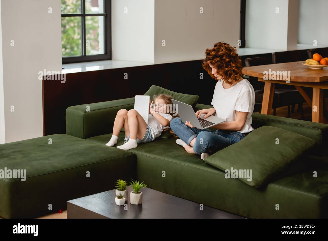 redhead freelancer using laptop near cute daughter with digital tablet Stock Photo