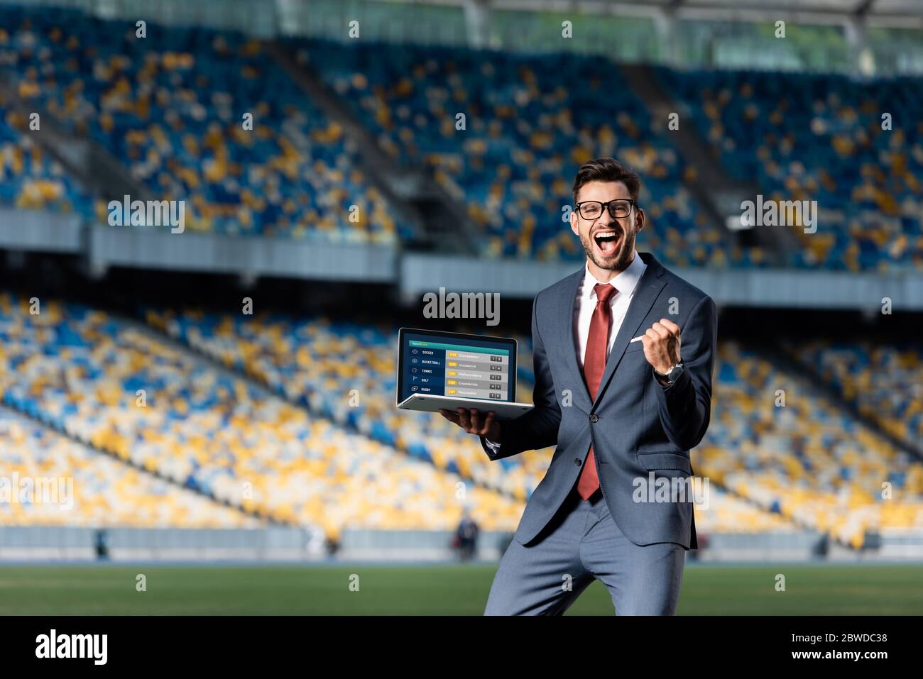 happy young businessman in suit holding laptop with sport betting website and showing yes gesture at stadium Stock Photo