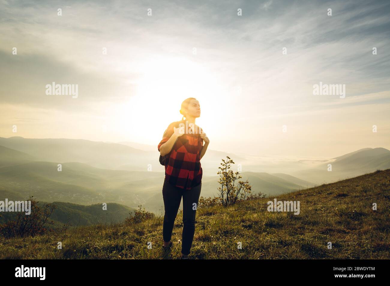 Young Woman Traveler In Red Plaid Shirt With Backpack Climbs Uphill At Sunrise. Scout Travel Adventure Concept Stock Photo