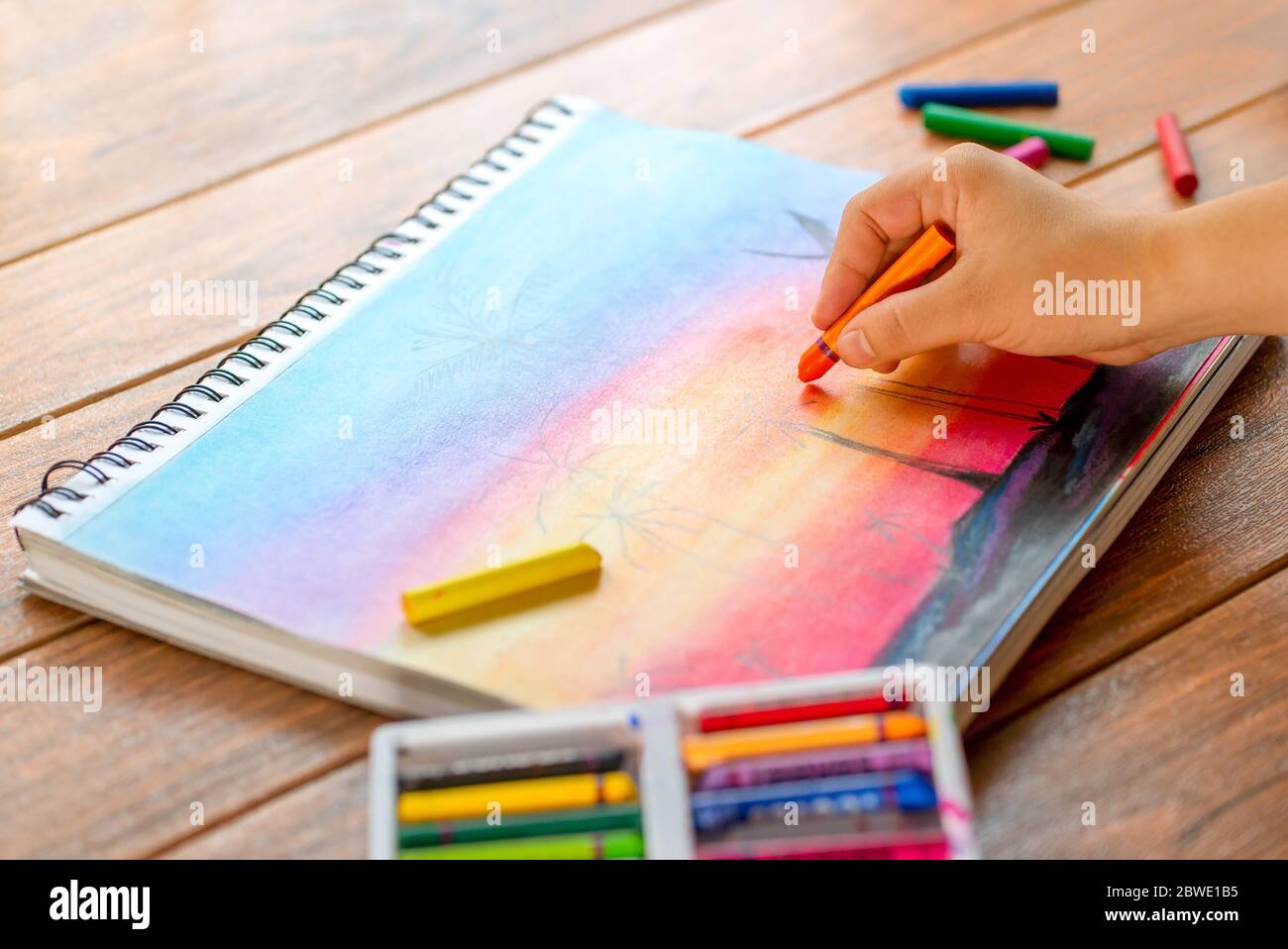 https://c8.alamy.com/comp/2BWE1B5/girls-hand-with-a-orange-wax-crayon-painting-on-a-white-paper-and-colorful-wax-crayons-in-the-background-2BWE1B5.jpg