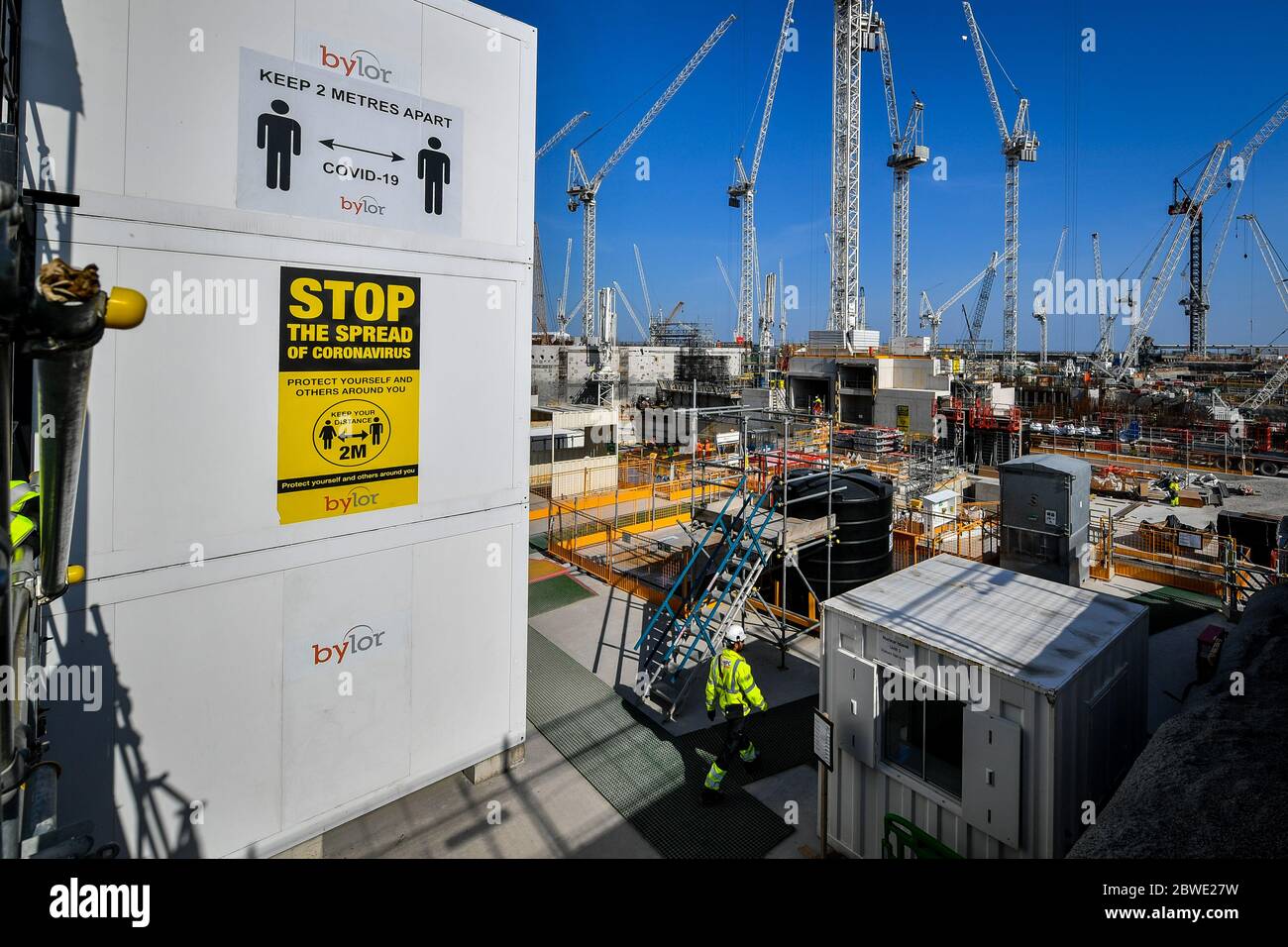 A construction worker passes below coronavirus advice signs at Hinkley Point C nuclear power station near Bridgwater, Somerset, Europe's largest building site, where they are completing a concrete pour into the base of unit 2 nuclear reactor. Stock Photo