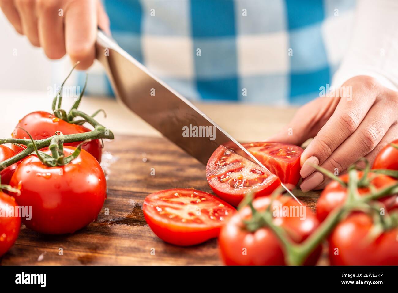 Detail of female hands cutting a tomato by a knife on a chopping board with more fresh tomatoes around Stock Photo