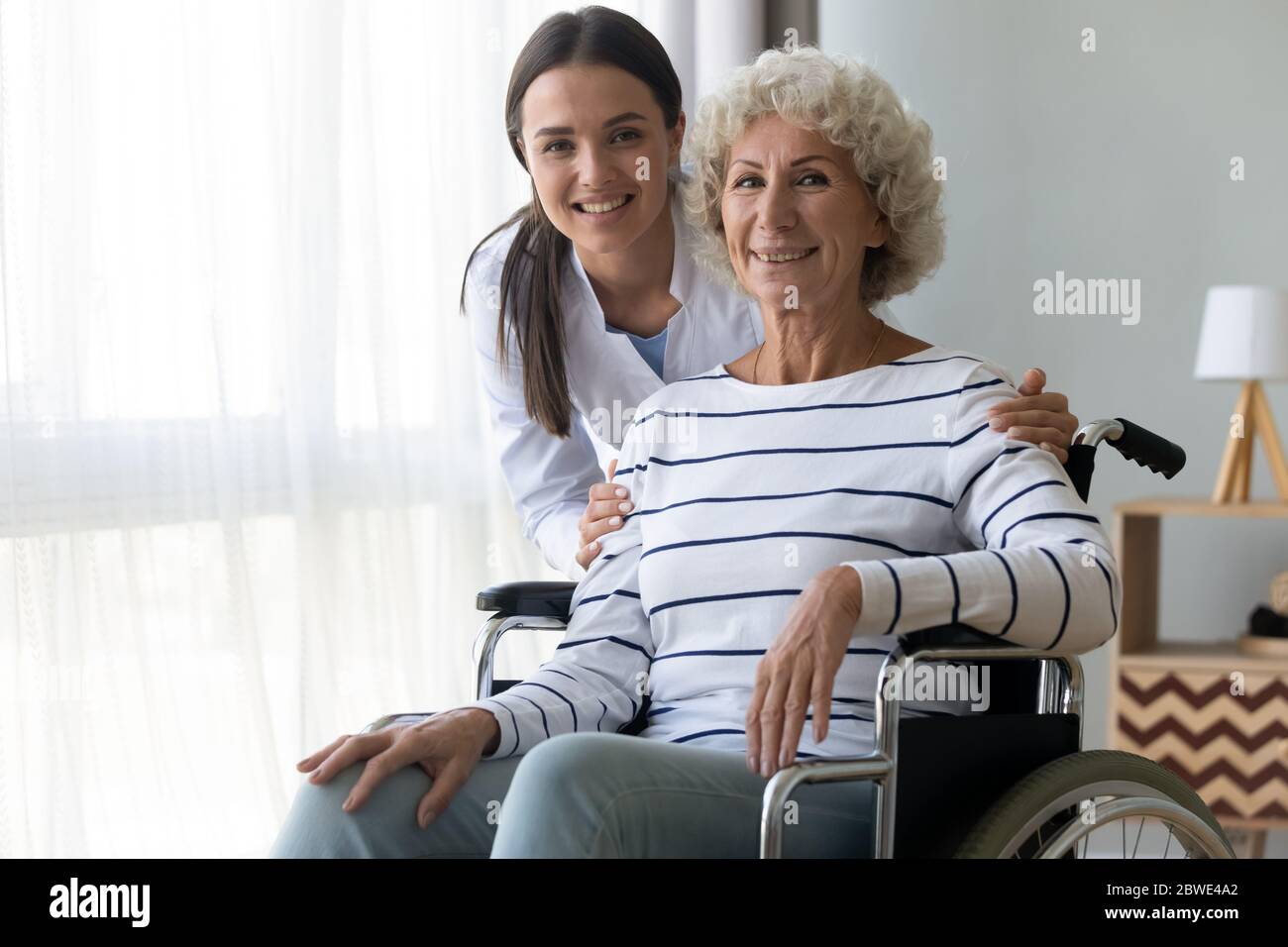 Caring nurse hug elderly disabled woman in wheelchair posing indoors Stock Photo