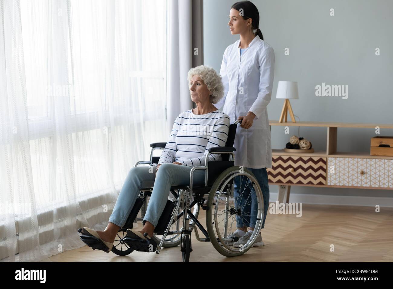Caring nurse and sad handicapped elderly woman in wheelchair indoors Stock Photo