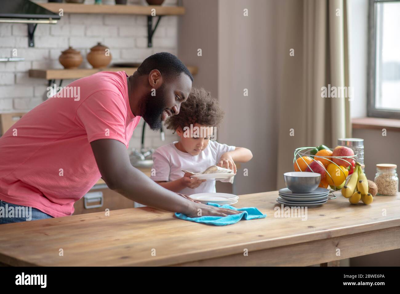 Tall dark-skinned man in a pink tshirt wiping the table and smiling, his daughter wiping the plate Stock Photo