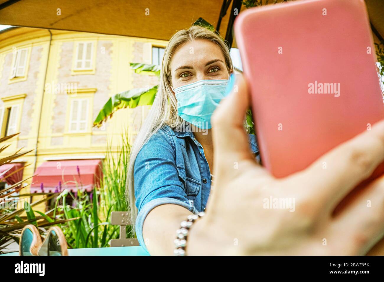 Girl in a garden bar taking a selfie with her face mask on to be protected from the coronavirus - Student holding smartphone in a videocall outdoors i Stock Photo