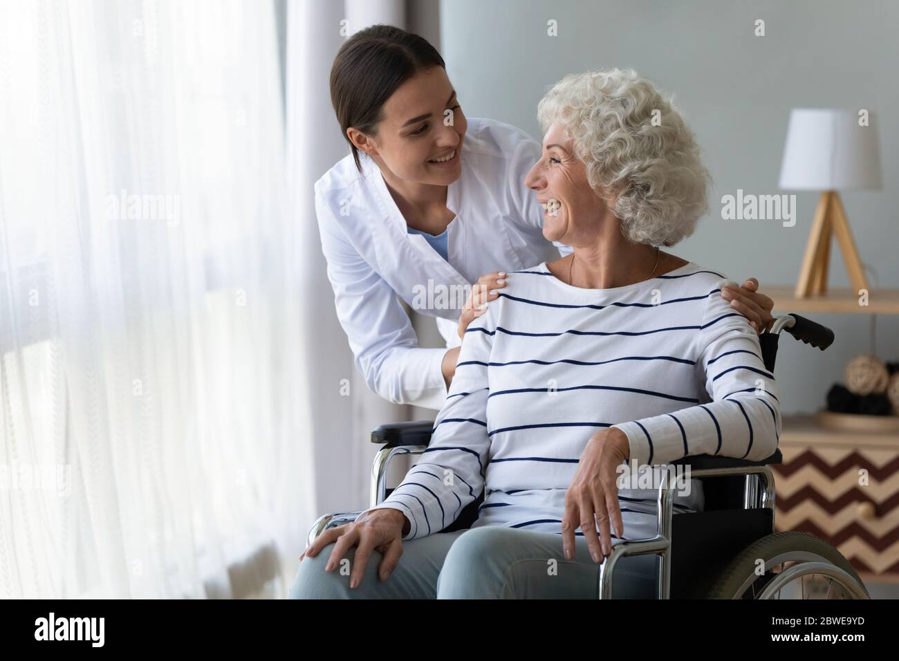 Smiling disabled elderly woman in wheelchair talking with caring nurse Stock Photo