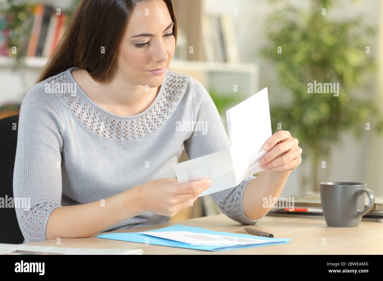 Serious woman opening envelope with letter inside sitting on a desk at home Stock Photo