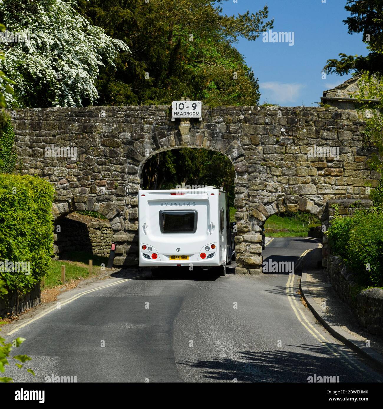 Caravan passing through narrow rustic stone archway spanning scenic country lane (tight squeeze) - B6160, Bolton Abbey village, Yorkshire, England, UK Stock Photo