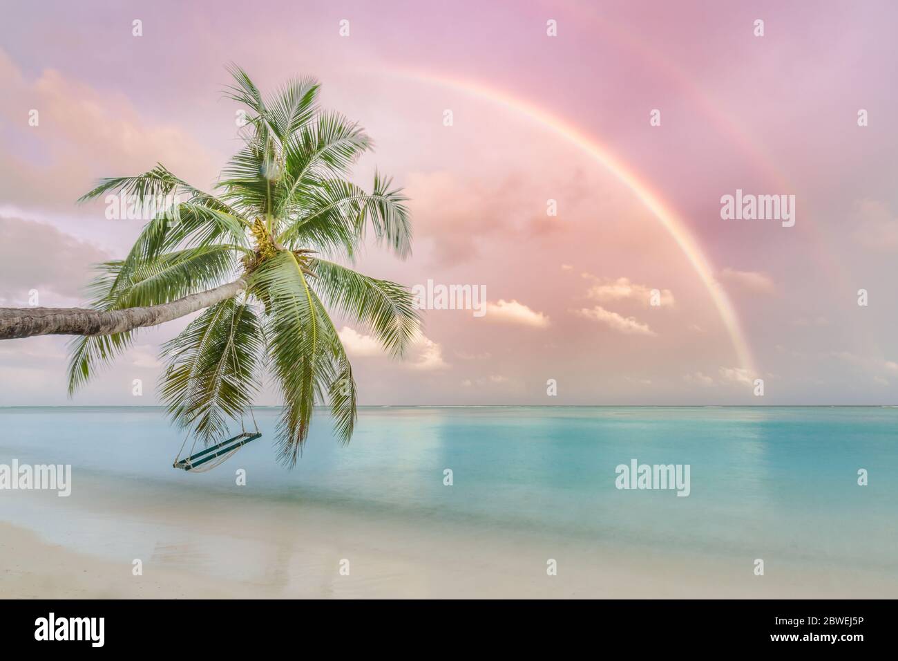 Perfect tropical landscape, sunset beach with palm tree and swing hanging under colorful rainbow. Romantic beach landscape Stock Photo