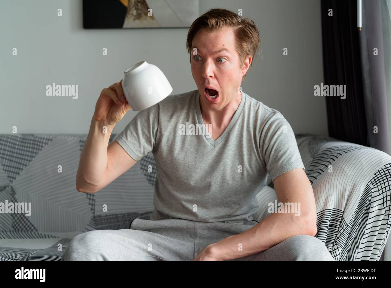 Young man looking shocked while holding coffee cup upside down at home