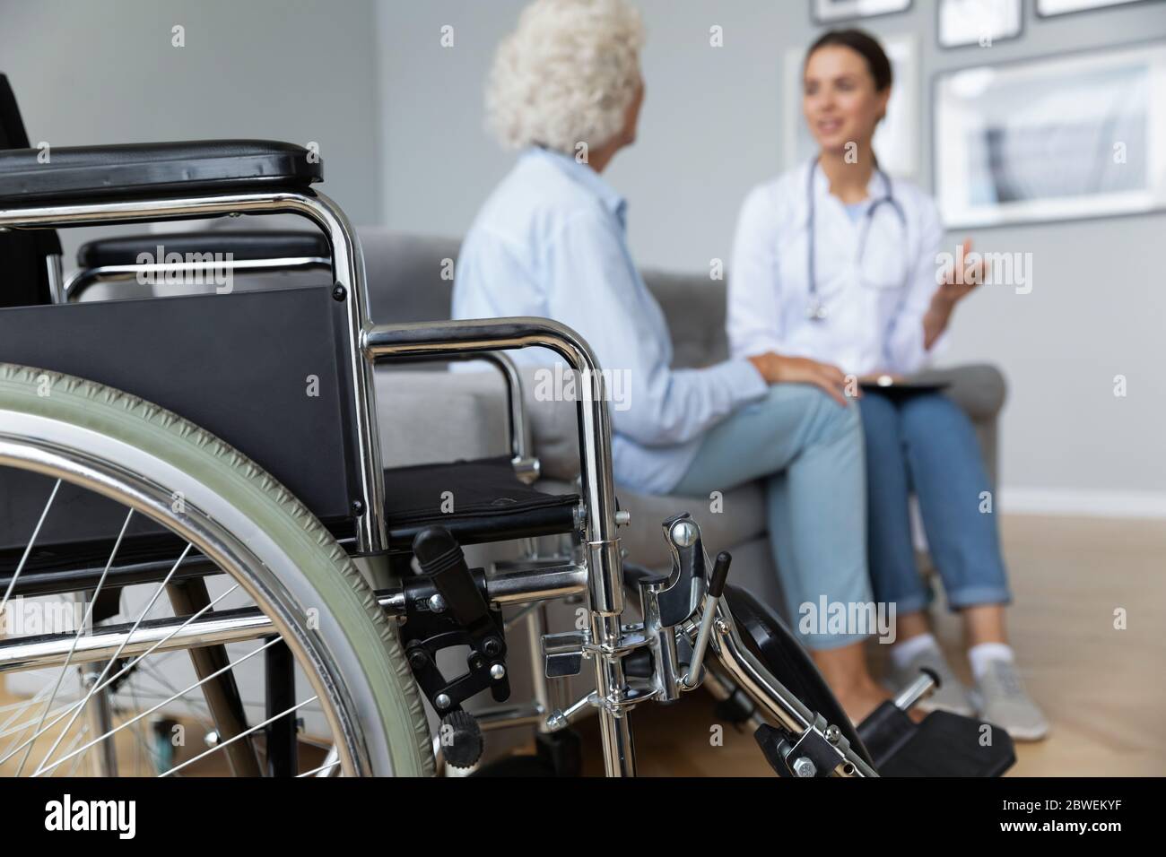 Physiotherapist elderly woman talking seated on sofa, focus on wheelchair Stock Photo