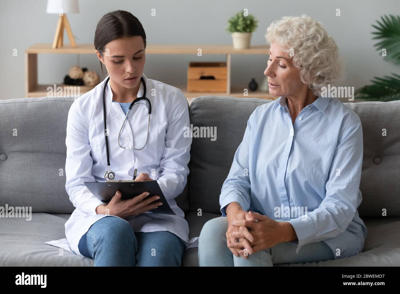 Young female caregiver listen writing health complaints of elderly woman Stock Photo