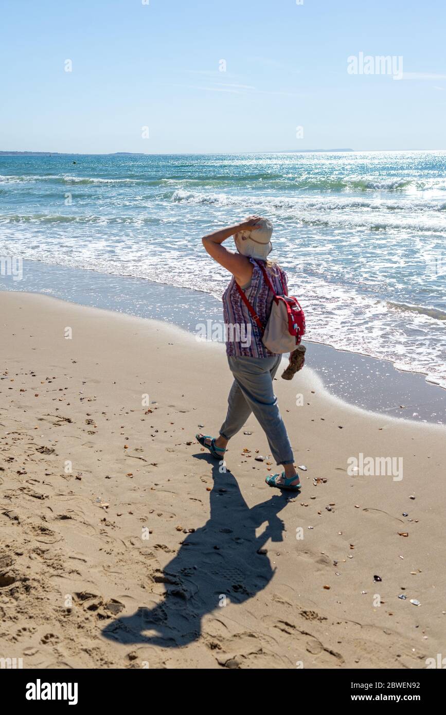 Branksome Chine Beach, Poole, Dorset, UK, 1st June 2020, Weather. More brilliant morning sunshine on the first day of meteorological summer following the sunniest spring on record in the UK. It’s Monday morning and the weekend crowds have gone. A woman walks along the shoreline holding onto her hat in the breeze. Credit: Paul Biggins/Alamy Live News Stock Photo