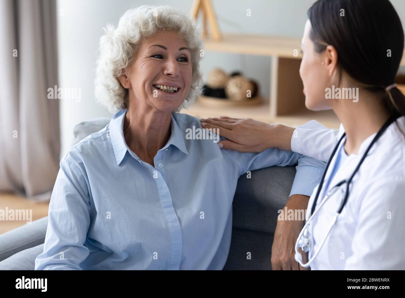 Elderly woman patient talking with young caregiver seated on couch Stock Photo