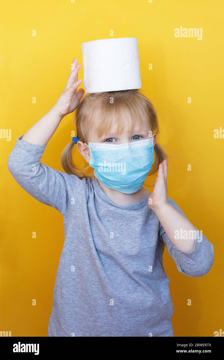 Cute little girl wearing a protective mask holding toilet paper roll on her head. Funny, positive photo. Corona virus scare. Epidemic Precautions. The Stock Photo