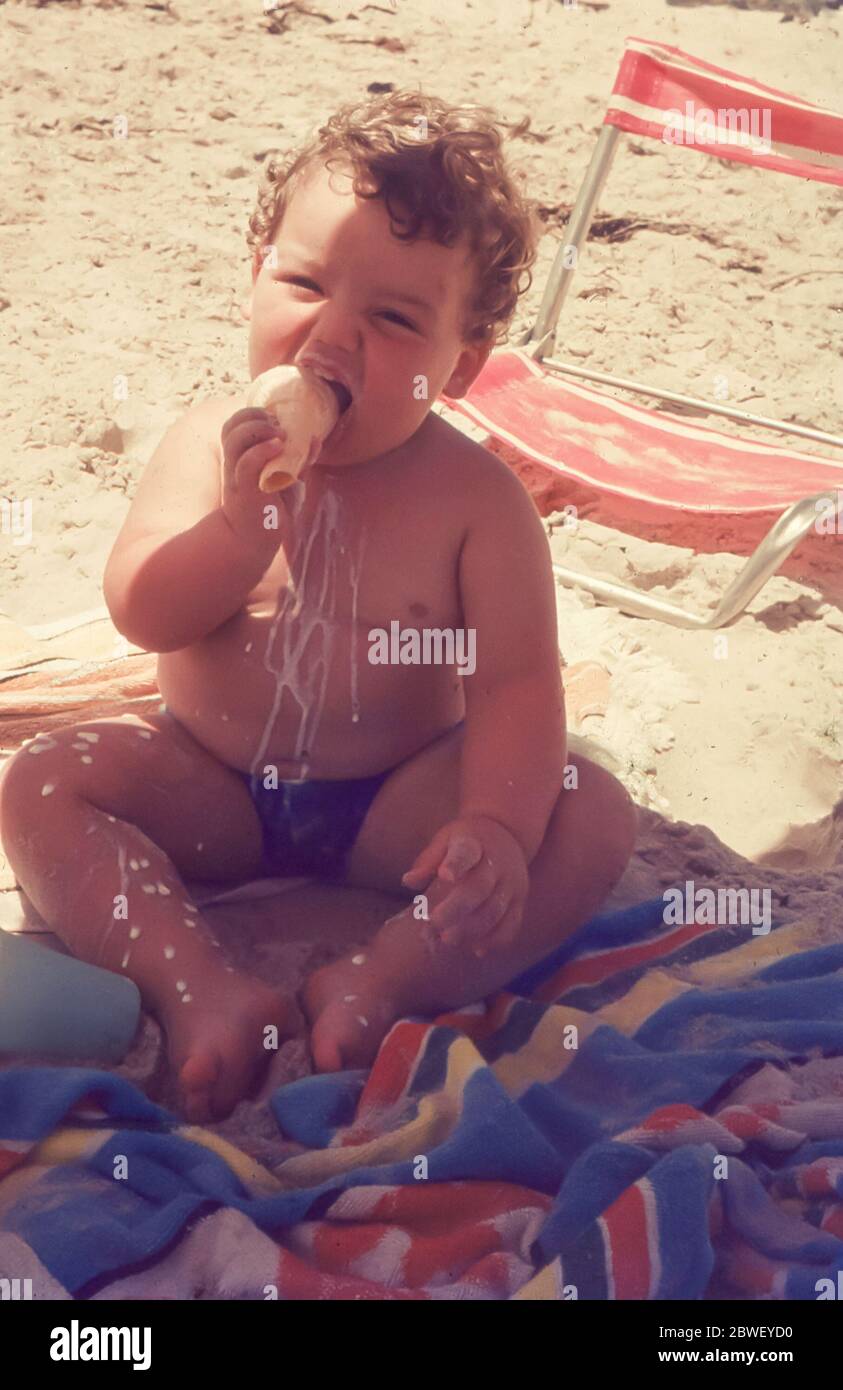 Sydney, Australia 1969: A young chubby curly haired Caucasian, 2 year old  boy sits on a beach under an umbrella eating an ice cream and spilling it  all down his front in