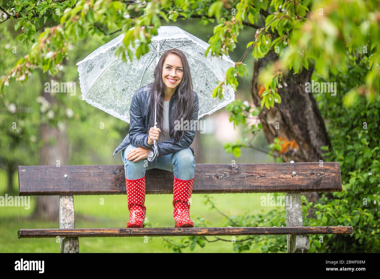 Smiling pretty woman holding umbrella on her shoulder sits on a bench back-rest in the park in the rain. Stock Photo