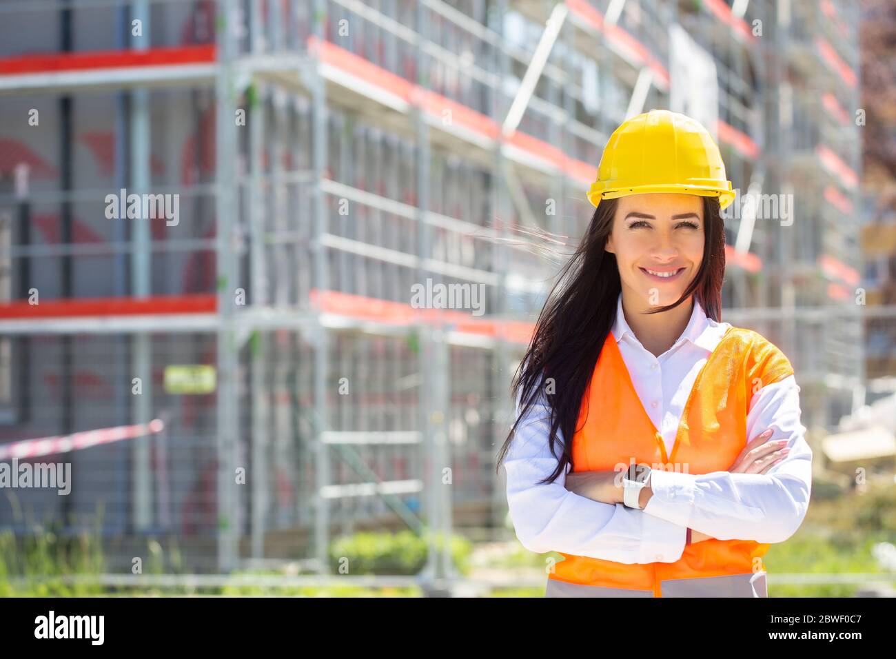 Female site supervisor wearing safety vest and helmet stands confidently in front of the construction covered in scaffolding. Stock Photo