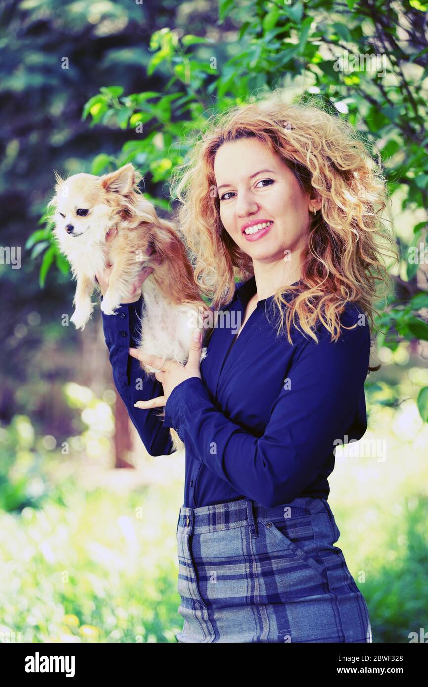 Blond young woman, business dressed, holding a long-haired chihuahua dog outdoor, on a green natural background ( with artistic filter ) Stock Photo