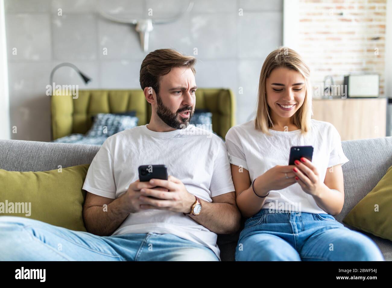 Young couple using smartphones at home. Jealous boyfriend spying his girlfriend's phone. Stock Photo