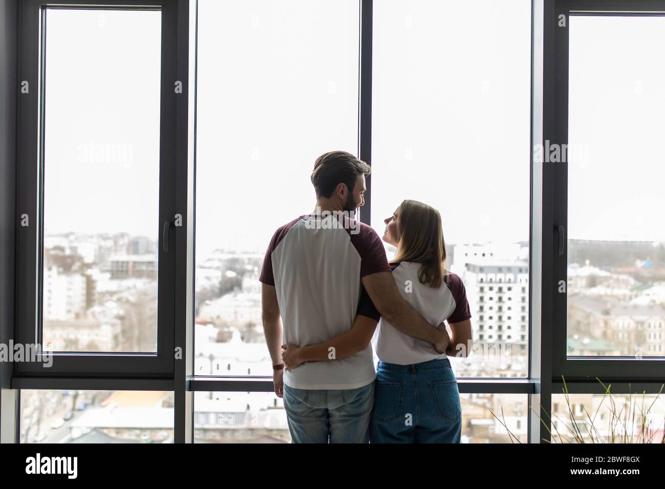 Young couple embracing standing near window and enjoying view from new apartment Stock Photo
