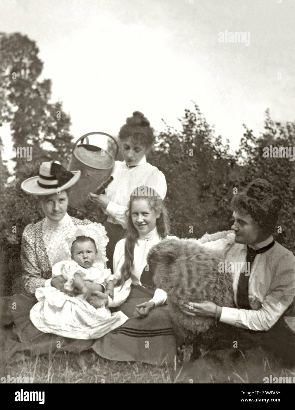 Four women (and surprisingly a sheep) find an amusing way of wetting the baby's head using a watering can on a Gloucestershire farm, England, UK c. 1900. To 'wet the baby's head' is a term used to celebrate the birth of a baby. Traditionally taking its name from the ceremony of a Christian baptism, during which the head of a baby would be anointed with holy water, the phrase now commonly relates to the general celebration of the new arrival. Stock Photo