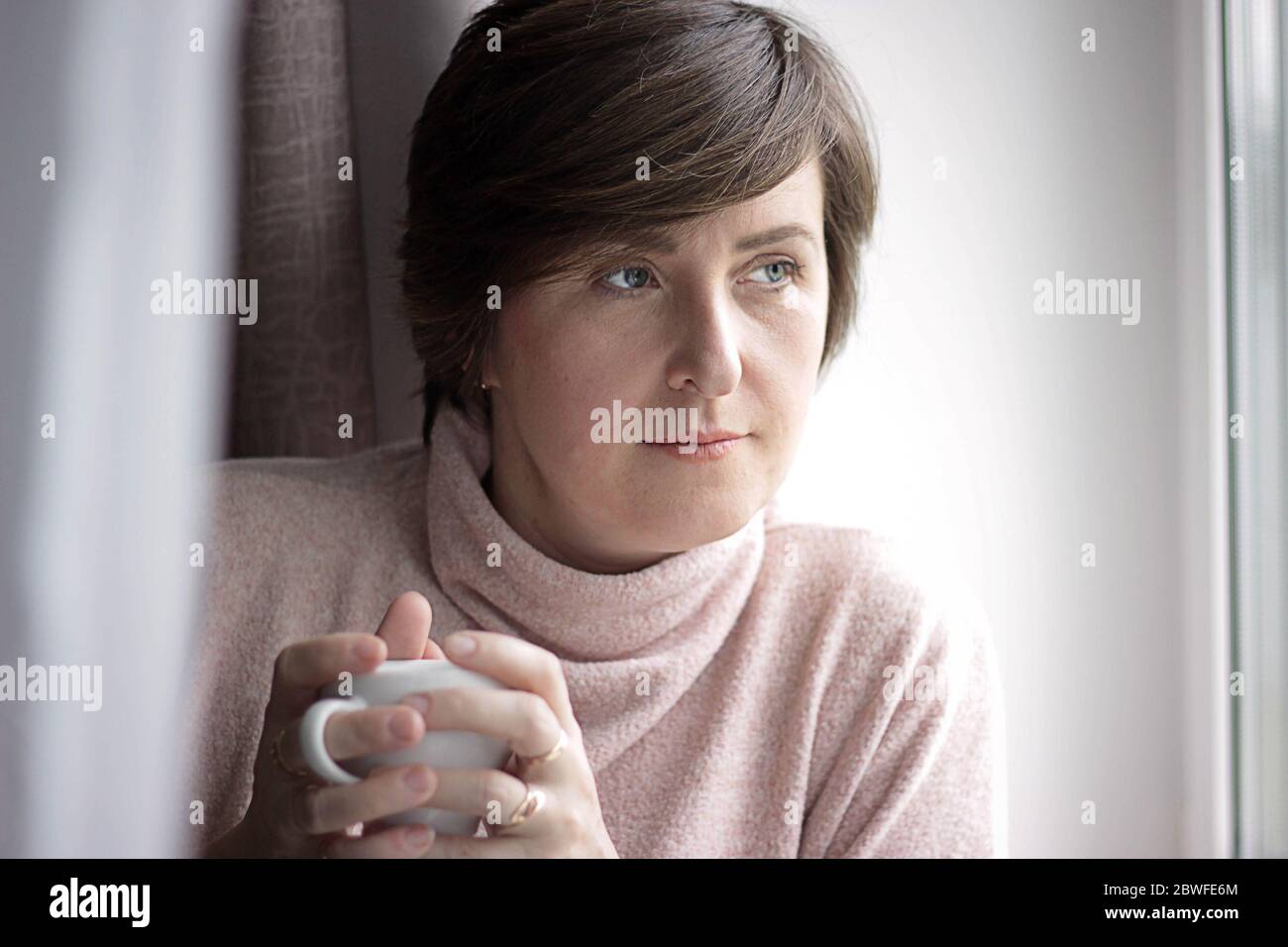 Girl at the window with a cup in winter. Home, sweater. Young Woman Enjoying her morning coffee or tea. Stock Photo