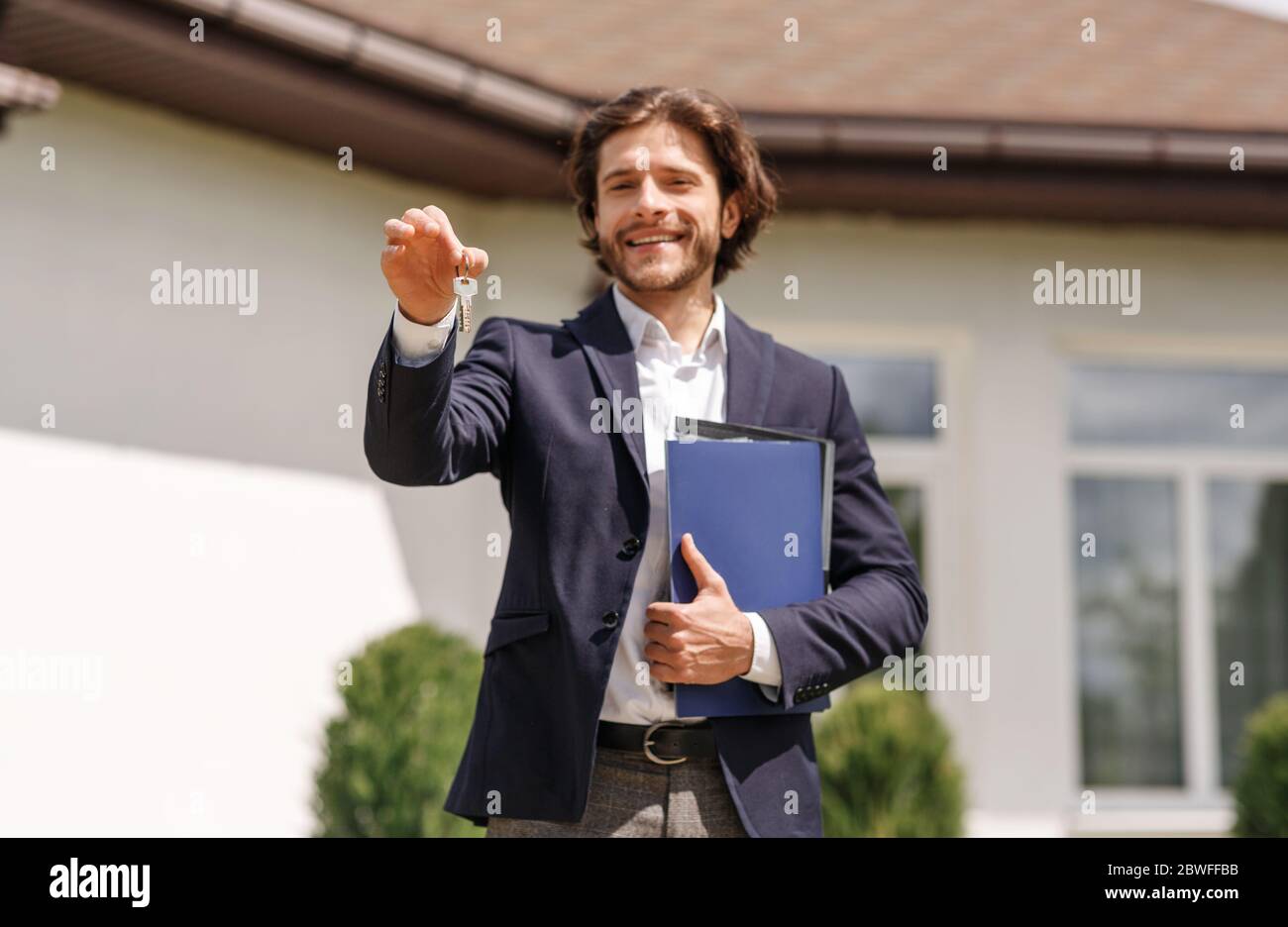 Smiling real estate agent holding house key near new residence to sale, outdoors Stock Photo