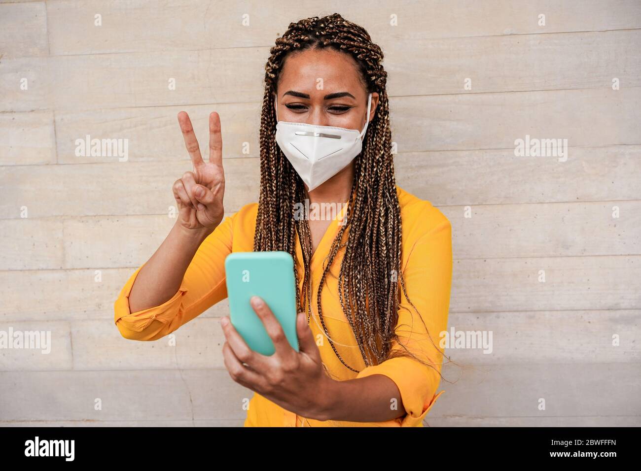 Young woman with braids doing video call while wearing face protective mask for coronavirus prevention - Happy girl streaming online outdoor during co Stock Photo