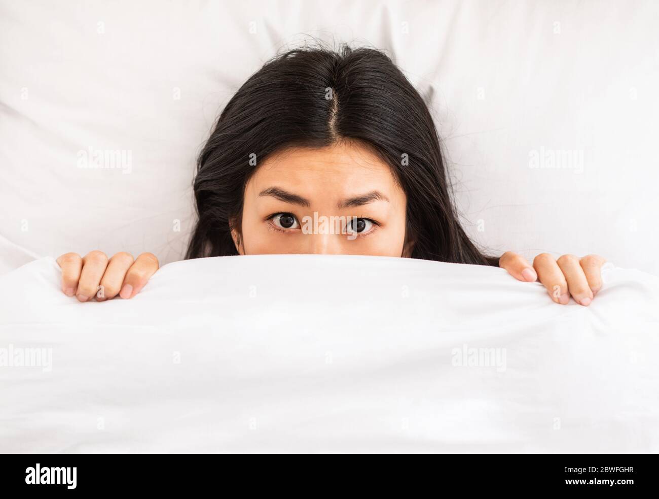 Chinese Girl Peeking Out Of Blanket Lying In Bed, Above-View Stock Photo